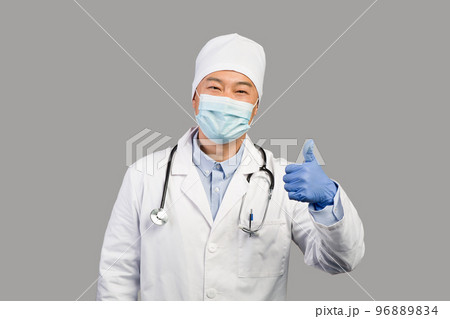 Smiling middle aged chinese male therapist in white coat, protective mask with stethoscope showing thumb up 96889834