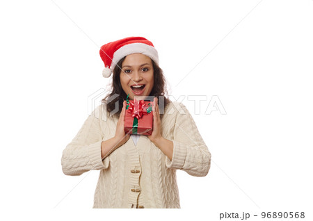 Adorable woman in Santa hat, holding Christmas present, expressing happiness, surprise, astonishment and positive emotions, smiling a toothy smile looking at camera, isolated over white background 96890568