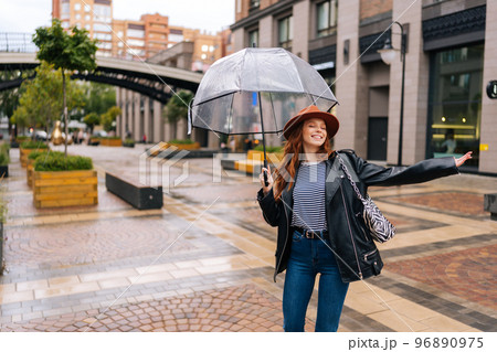 Cheerful excited young woman in elegant hat dancing and having fun with transparent umbrella on beautiful city street, enjoying rainy weather outdoors. Concept of female lifestyle at autumn season. Cheerful excited young woman in elegant hat dancing and having fun with transparent umbrella on beautiful city street, enjoying rainy weather outdoors. Concept of female lifestyle at autumn season. 96890975