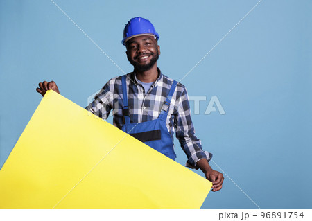 Relaxed construction worker with protective helmet presenting a panel with advertising space. Builder with overalls holding empty yellow banner for construction industry concept on blue background. 96891754