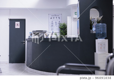 Corridor of waiting area of medical clinic with front desk for patient care and office furniture. No one at empty counter in medical office lobby seeing patients. Clinic with no doctors or sick people Corridor of waiting area of medical clinic with front desk for patient care and office furniture. No one at empty counter in medical office lobby seeing patients. Clinic with no doctors or sick people 96891838