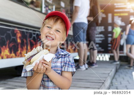 Cute adorable little happy smiling boy kid enjoy eating hot dog sausage in bread near street cafe stall outdoors. Child healthy eating lunch hotdog. Junk food and fast food unhealthy snack concept Cute adorable little happy smiling boy kid enjoy eating hot dog sausage in bread near street cafe stall outdoors. Child healthy eating lunch hotdog. Junk food and fast food unhealthy snack concept 96892054