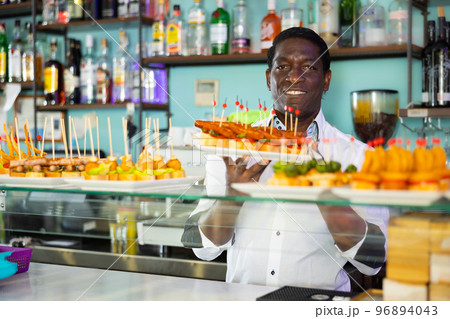 Smiling African American bartender standing with plates of pinchos in front of a pub counter 96894043