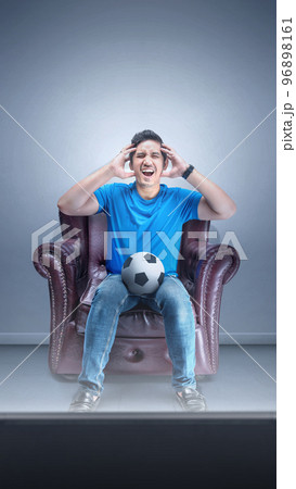 Asian man with the ball sitting on the couch with a disappointed expression watching football 96898161
