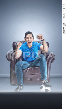 Asian man with the ball sitting on the couch with an excited expression watching football 96898163