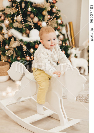 A blonde girl in a sweater rides a wooden horse at home against the background of the Christmas tree. Cute little girl playing at home on the background of garlands. 96907485