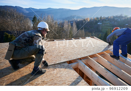 Carpenters mounting wooden OSB board on rooftop of future cottage. Men workers building wooden frame house. Carpentry and construction concept. Carpenters mounting wooden OSB board on rooftop of future cottage. Men workers building wooden frame house. Carpentry and construction concept. 96912209
