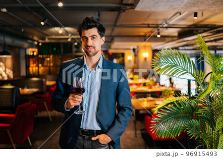 Portrait of confident elegant man in fashion suit holding glasses of red wine standing in restaurant with luxury interior, smiling looking at camera. Positive bearded male owner welcoming guests. 96915493