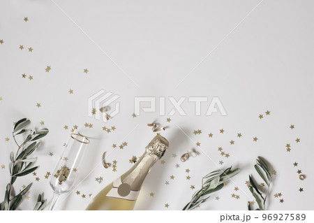 New Year festive web banner. Birthday, wedding party. Celebration concept. Champagne wine bottle, drinking glass. Olive tree branches. White table background. Golden star confetti. Flat lay, top view. 96927589