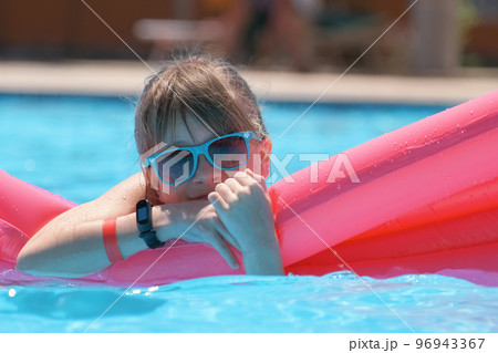 Young joyful child girl having fun swimming on inflatable air mattress in swimming pool with blue water on warm summer day on tropical vacations. Summertime activities concept Young joyful child girl having fun swimming on inflatable air mattress in swimming pool with blue water on warm summer day on tropical vacations. Summertime activities concept 96943367
