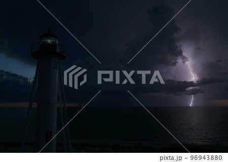 White tall lighthouse on sea shore with blinking light at stormy night for commercial vessels navigation. Thunderstorm with lightnings over ocean water posing danger for ships White tall lighthouse on sea shore with blinking light at stormy night for commercial vessels navigation. Thunderstorm with lightnings over ocean water posing danger for ships 96943880