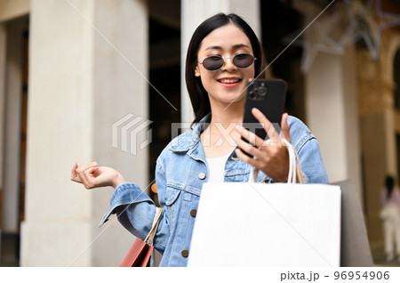 Joyful Asian female shopper using her smartphone while enjoys shopping in the mall. 96954906