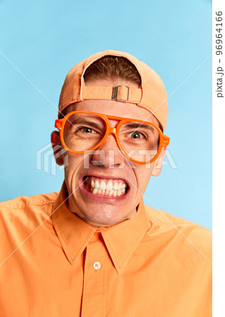 Closeup portrait of emotional young man in eyewear and cap looking at camera with vivid emotions isolated over light blue background. Mental health, facial expression, emotions concept Closeup portrait of emotional young man in eyewear and cap looking at camera with vivid emotions isolated over light blue background. Mental health, facial expression, emotions concept 96964166