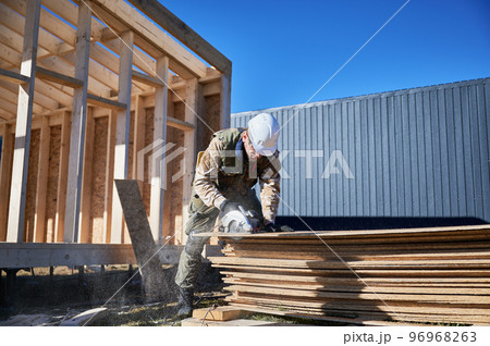 Carpenter using circular saw for cutting wooden OSB board. Man worker building wooden frame house on pile foundation. Carpentry concept. 96968263