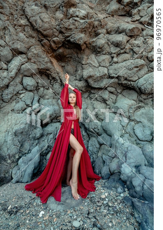 Red dress rocks woman. A blonde with flowing hair in a long flowing red dress stands near a rock of volcanic origin. Travel concept, photo session at sea 96970565