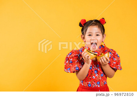 Chinese New Year. Happy Asian Chinese little girl smile wearing red cheongsam holding gold ingot, Portrait children in traditional dress hold golden bar, studio short isolated on yellow background Chinese New Year. Happy Asian Chinese little girl smile wearing red cheongsam holding gold ingot, Portrait children in traditional dress hold golden bar, studio short isolated on yellow background 96972810