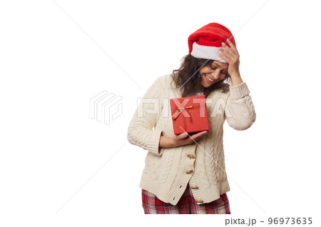 Waist-up portrait of a beautiful multi-ethnic positive woman, wearing Santa hat and warm knitted sweater, rejoicing at happy Christmas present, isolated over white background. Copy advertising space 96973635
