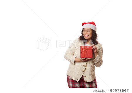 Waist-up portrait on white background of a multi-ethnic positive pretty woman wearing Santa hat, cheerfully smiling looking aside at copy ad space, posing with gift box with Christmas happy present 96973647
