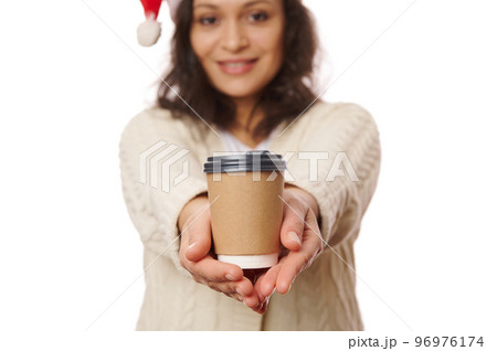 Selective focus on a takeaway hot drink in disposable cardboard cup in the hands of a blurred woman in Santa hat, smiling on camera, isolated over white background. Beverage to go. Mockup. Copy space. Selective focus on a takeaway hot drink in disposable cardboard cup in the hands of a blurred woman in Santa hat, smiling on camera, isolated over white background. Beverage to go. Mockup. Copy space. 96976174