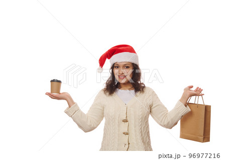 Isolated portrait on white background of a pretty woman in Santa hat and beige warm sweater, smiling on camera while holding disposable cup of takeaway hot coffee or tea and paper bag. Beverage to go 96977216