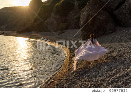 A mysterious female silhouette with long braids stands on the sea beach with mountain views, Sunset rays shine on a woman. Throws up a long white dress, a divine sunset. 96977632