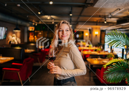 Portrait of attractive female holding glass of red wine standing in restaurant with luxury dark interior, looking away. Front view of cute woman posing at cafe, blurred background. Portrait of attractive female holding glass of red wine standing in restaurant with luxury dark interior, looking away. Front view of cute woman posing at cafe, blurred background. 96978929