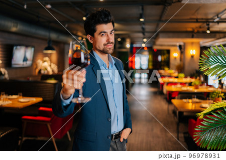 Portrait of smiling elegant man in fashion suit holding glasses of red wine standing in restaurant with luxury interior, looking at camera. Successful bearded male owner welcoming guests. Portrait of smiling elegant man in fashion suit holding glasses of red wine standing in restaurant with luxury interior, looking at camera. Successful bearded male owner welcoming guests. 96978931