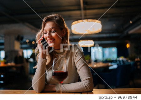 Portrait of cheerful blonde female talking on mobile phone sitting at table with red wine in modern restaurant with dark interior. Attractive happy young woman having conversation on smartphone in bar 96978944
