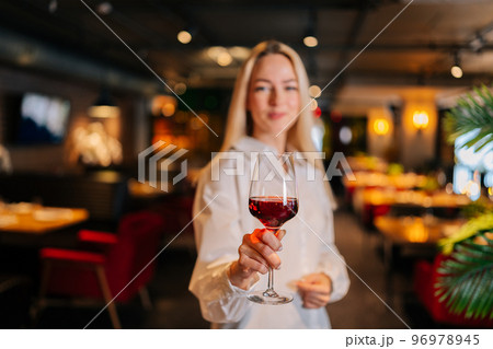 Selective focus portrait of blonde woman holding glass of red wine standing in restaurant with luxury interior, looking at camera. Front view of cute female posing at cafe, blurred background 96978945