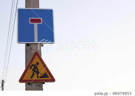 Bright yellow triangular warning sign; Attention, work ahead, and Dead-end street on square blue shield on concrete pillar on clear blue sky copy space background. Repair, road construction concept. Bright yellow triangular warning sign; Attention, work ahead, and Dead-end street on square blue shield on concrete pillar on clear blue sky copy space background. Repair, road construction concept. 96979953