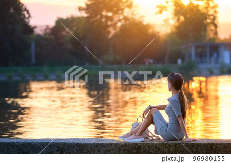 Back view of lonely young woman sitting alone on lake shore enjoying warm evening. Wellbeing and relaxing in nature concept. 96980155