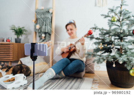 Focus on phone with young Woman playing ukulele guitar and recording Christmas music video for social networks on phone in modern Scandi interior home. Musician blogger singing song, content creating. 96980192