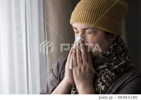 Sick brunette woman in cap and scarf sneezes covering nose with napkin feeling unwell. Sick brunette woman in cap and scarf sneezes covering nose with napkin feeling unwell. 96983280
