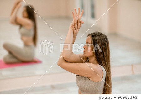 Young woman doing yoga in the gym. A girl with long hair and in a beige tracksuit stands in a cow pose on a pink carpet. A woman performs Gomukhasana. Young woman doing yoga in the gym. A girl with long hair and in a beige tracksuit stands in a cow pose on a pink carpet. A woman performs Gomukhasana. 96983349