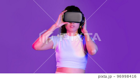Young asian woman in white t-shirt wearing vr headset looking playing touching purple screen color background. Metaverse concept. Young asian woman in white t-shirt wearing vr headset looking playing touching purple screen color background. Metaverse concept. 96998416