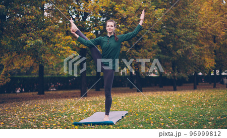 Smiling girl professional yoga instructor is doing balancing exercises standing on one leg on mat on green and yellow grass in park. Beautiful autumn nature is visible. Smiling girl professional yoga instructor is doing balancing exercises standing on one leg on mat on green and yellow grass in park. Beautiful autumn nature is visible. 96999812