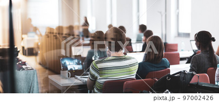 Workshop at university. Rear, trough the window, view of students sitting and listening in lecture hall doing practical tasks on their laptops. 97002426