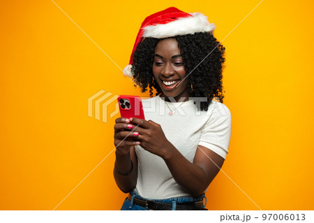 Attractive african lady in Santa hat using her cellphone on yellow background Attractive african lady in Santa hat using her cellphone on yellow background 97006013