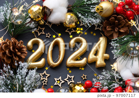 The golden figures 2024 made of candles on a black stone slate background are decorated with a festive decor of stars, sequins, fir branches, balls and garlands. Greeting card, happy New Year. 97007117