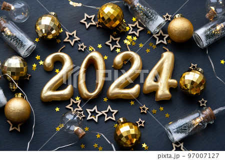 The golden figures 2024 made of candles on a black stone slate background are decorated with a festive decor of stars, sequins, fir branches, balls and garlands. Greeting card, happy New Year. 97007127