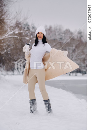 A beautiful girl in a beige cardigan and a white hat with a glass of tea enjoys a snowy embankment by the lake A beautiful girl in a beige cardigan and a white hat with a glass of tea enjoys a snowy embankment by the lake 97007942