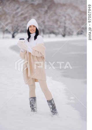 A beautiful girl in a beige cardigan and a white hat with a glass of tea enjoys a snowy embankment by the lake 97007949