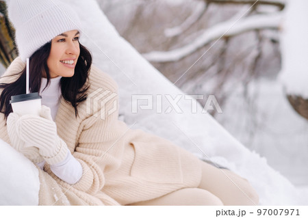 A beautiful girl with a beige cardigan and a white hat enjoying drinking tea in a snowy winter forest near a lake A beautiful girl with a beige cardigan and a white hat enjoying drinking tea in a snowy winter forest near a lake 97007971