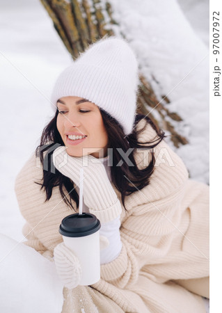A beautiful girl with a beige cardigan and a white hat enjoying drinking tea in a snowy winter forest near a lake 97007972