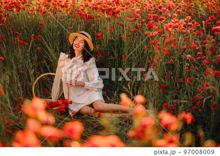 A girl in a white dress and with a basket of poppies is sitting on a poppy field 97008009