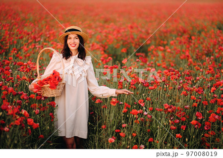 A girl in a white dress and with a basket of poppies walks through a poppy field 97008019