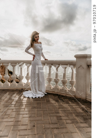 Romantic bride, blond girl in white wedding dress with open shoulders posing on open terrace with backdrop of the sea and rocks. Stylish young woman standing on terrace and looking on ocean at sunset 97010719