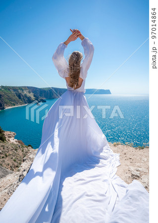 Blonde with long hair on a sunny seashore in a white flowing dress, rear view, silk fabric waving in the wind. Against the backdrop of the blue sky and mountains on the seashore. Blonde with long hair on a sunny seashore in a white flowing dress, rear view, silk fabric waving in the wind. Against the backdrop of the blue sky and mountains on the seashore. 97014864