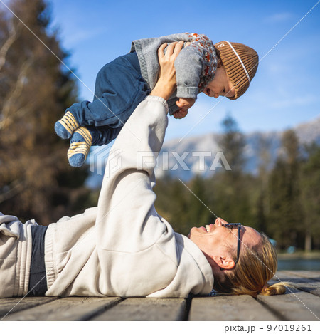 Happy family. Young mother playing with her baby boy infant oudoors on sunny autumn day. Portrait of mom and little son on wooden platform by lake. Positive human emotions, feelings, joy Happy family. Young mother playing with her baby boy infant oudoors on sunny autumn day. Portrait of mom and little son on wooden platform by lake. Positive human emotions, feelings, joy 97019261