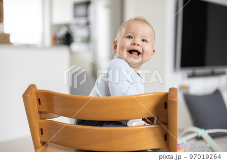 Happy infant sitting in traditional scandinavian designer wooden high chair and laughing out loud in modern bright home. Cute baby smile Happy infant sitting in traditional scandinavian designer wooden high chair and laughing out loud in modern bright home. Cute baby smile 97019264